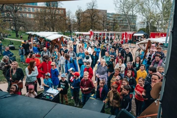 Pohled na zaplněné prostranství s návštěvníky festivalu, červené stánky v pozadí.