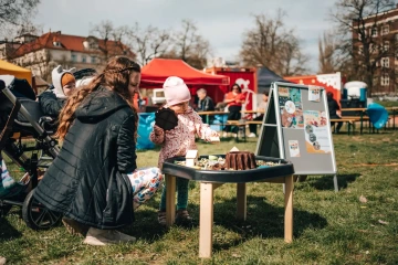Dětský koutek na festivalu, maminka s dítětem si hrají u stolečku s hračkami na trávě.