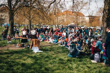 Návštěvníci sedí na dekách a sledují program na hlavním pódiu, kolem rozkvetlé stromy.