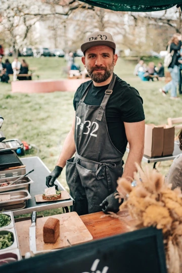 Muž v čepici a zástěře stojí u venkovního stánku s jídlem a usmívá se na návštěvníky.