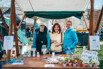 Stánek s květináči a produkty na podporu charitativní sbírky, skupina žen pózuje k fotografii.