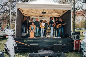 Hudební kapela na pódiu hraje pro publikum, vedle pódia stojí kreslená figurka symbolizující charitativní projekt.