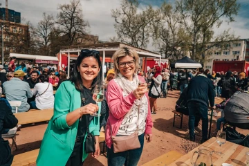 Dvě ženy pózují se skleničkami v ruce u piknikových stolů festivalu.