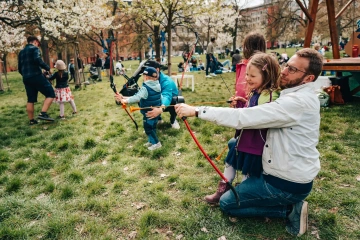 Dospělý muž pomáhá malé dívce s lukostřelbou na travnaté ploše festivalu.