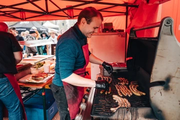 Muž v modrém tričku a zástěře griluje klobásy na velkém grilu pod červeným stanem během festivalu. V pozadí je vidět fronta návštěvníků čekajících na jídlo.