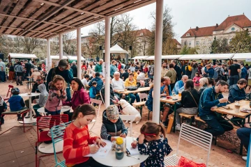 Velká krytá terasa plná návštěvníků sedících u stolů s jídlem a nápoji během festivalového dne.