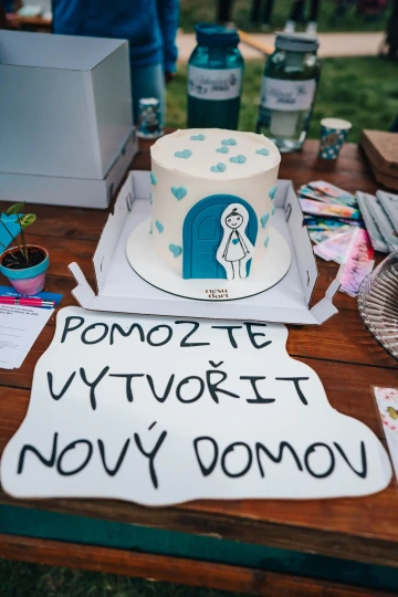 Detail dortu s kresbou dívky, která je maskotem Tady to mám rád, stojící před modrými dveřmi, vystaveného na charitativním stánku vedle pokladniček a letáků. Pod sebou má nápis Pomozte vytvořit Nový domov.