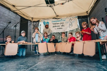 Skupina dětí sedí za stolem na pódiu, účastní se festivalového programu, v pozadí banner akce.