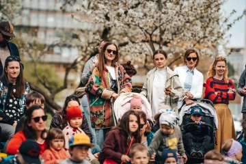 Skupina žen s dětmi a kočárky sleduje program festivalu v parku, kolem kvetoucí stromy.