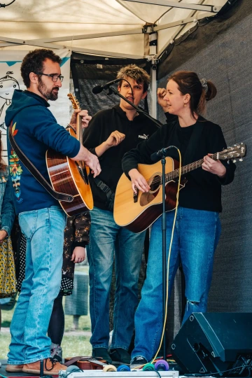 vě ženy a muž s kytarami připravují hudební vystoupení na pódiu festivalu.