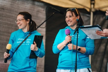 Dvě moderátorky v modrých tričkách s logem festivalu se usmívají a hovoří do mikrofonů na pódiu.