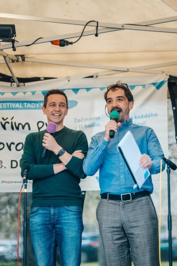 Dva moderátoři stojí na pódiu s mikrofony, smějí se a moderují program festivalu. Jeden z nich je Jan Grolich.