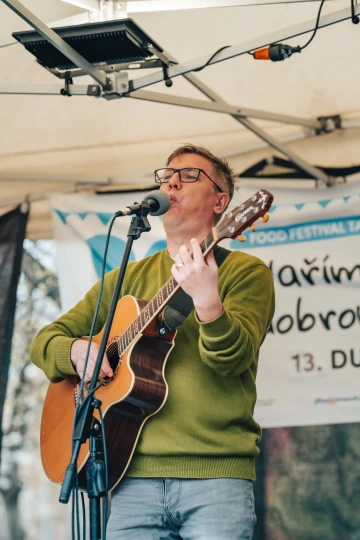 Muž ve svetru hraje na akustickou kytaru a zpívá na venkovním pódiu během festivalu.