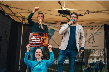 Dva moderátoři stojí na pódiu, jeden drží mikrofon a zvedá ruku, v pozadí je vidět banner festivalu s charitativním motivem.