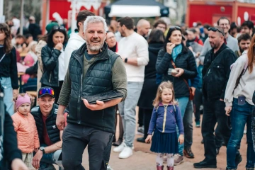 Muž s šedými vlasy a vousy drží v ruce talíř s jídlem a kráčí směrem k fotografovi, za ním je dav návštěvníků festivalu včetně dětí a rodin.