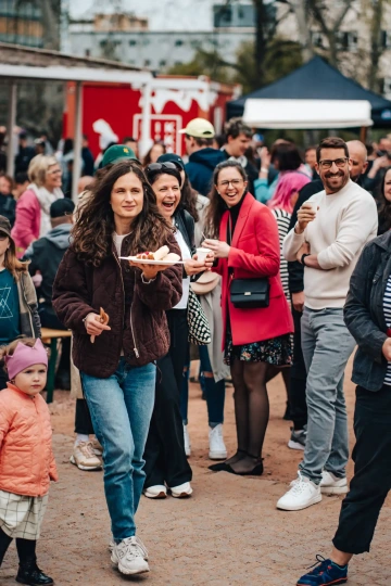 Skupina návštěvníků stojí ve frontě na občerstvení, v popředí ženy nesoucí talíře s jídlem.