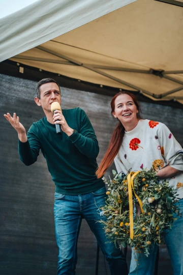 Muž s mikrofonem a žena s květinovým věncem a barevnými stuhami na pódiu mluví k návštěvníkům festivalu.