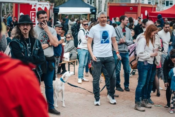 Návštěvníci různých věkových kategorií se procházejí kolem stánků, v popředí muž se psem.