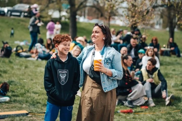 Žena a dospívající chlapec stojí na trávě mezi ostatními návštěvníky a smějí se, v ruce drží nápoje.