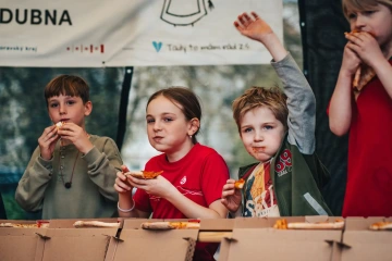 Tři děti u tvořivého stolu na festivalu, drží v rukou malé pečivo a sledují dění kolem sebe.