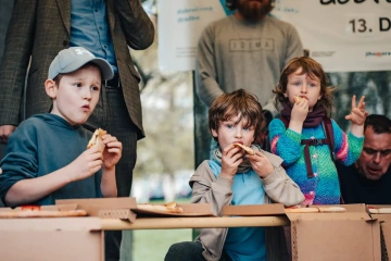 Dvě děti a dospělí sedí u stolu na festivalu, děti se soustředěně věnují tvoření a pojídají drobné občerstvení.
